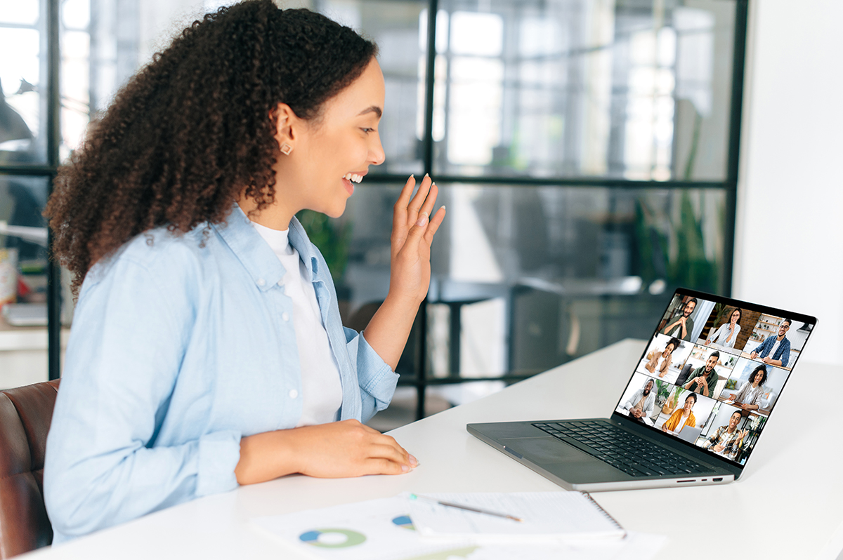 Side view of brazilian businesswoman, ceo, team leader, sitting at work dest in the office in front of a laptop, talking by online video conference with multiracial team, having brainstorm, smile